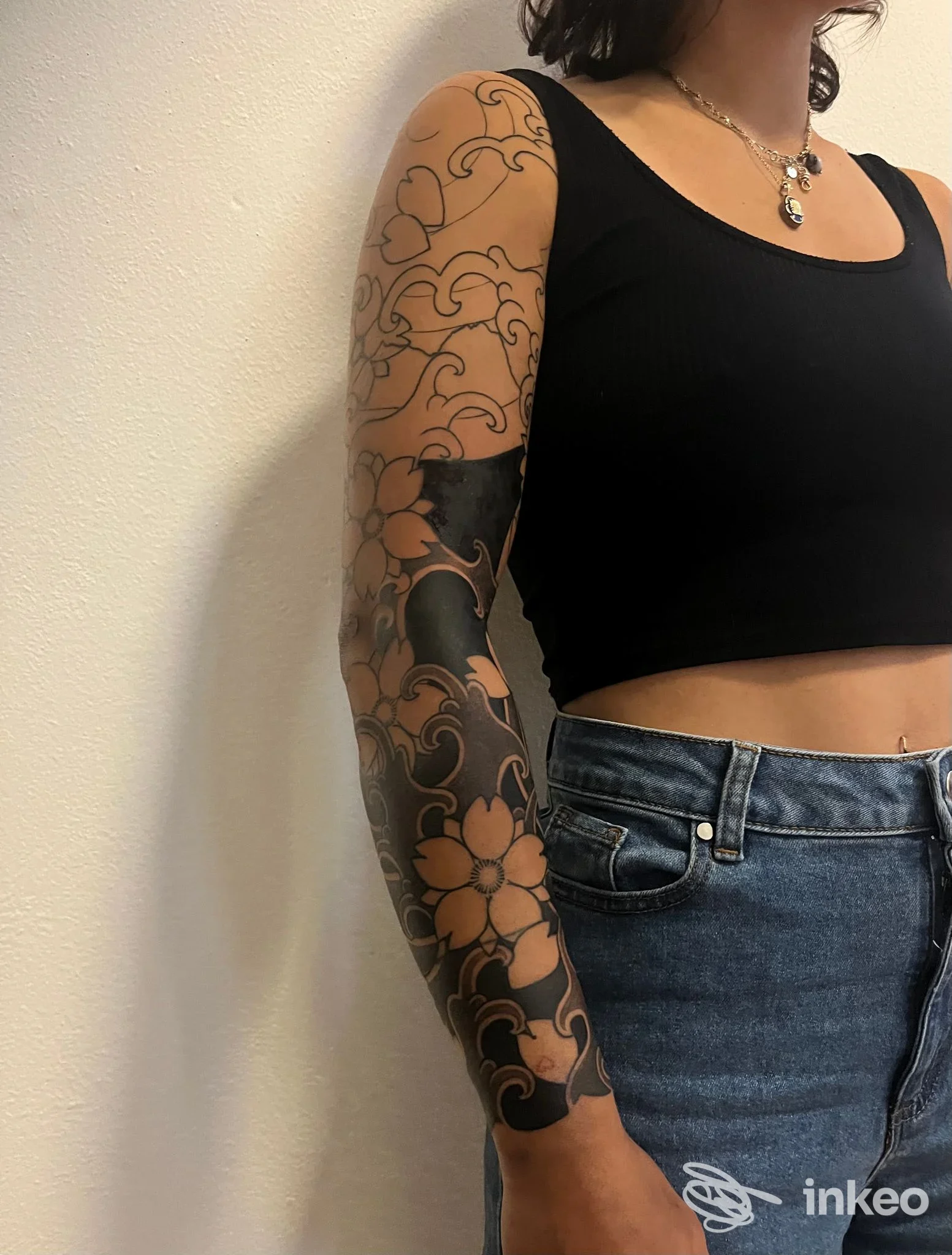 Braccio di una persona con tatuaggio lungo braccio stile floreale in bianco e nero, indosso maglietta nera crop e jeans, sfondo neutro. studio ĀYU Tattoo Temple, Cremona.