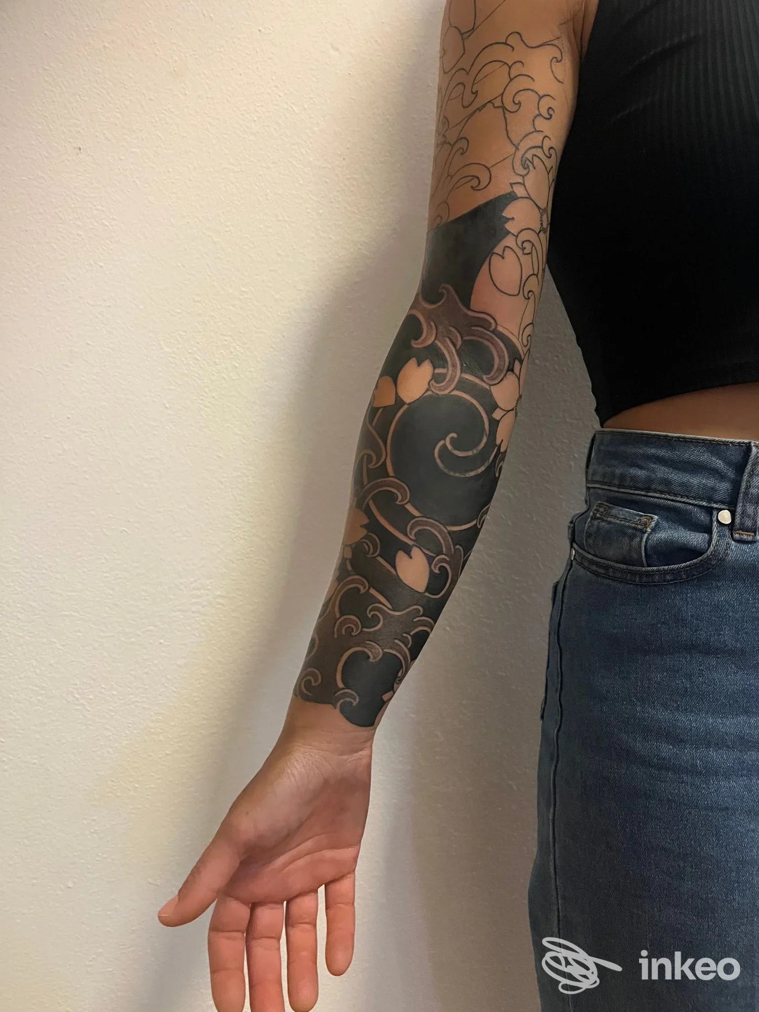 Braccio di persona con tatuaggio a braccia lunghe di motivo floreale scure su pelle, visibile da polso a metà avambraccio, vestito con manica corta nera e jeans.
