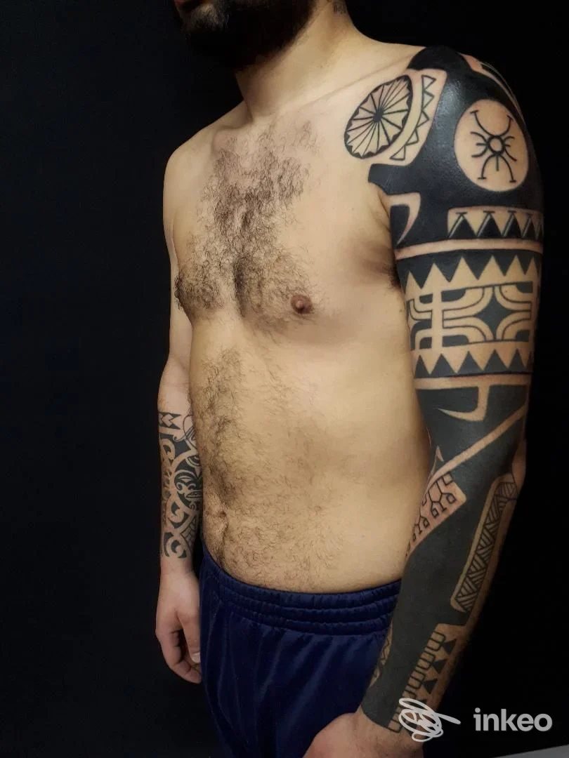 Uomo senza camicia con braccio destro tattooato in stile geometrico, braccio e petto visibili su sfondo scuro. Foto frontale con illuminazione diretta.
