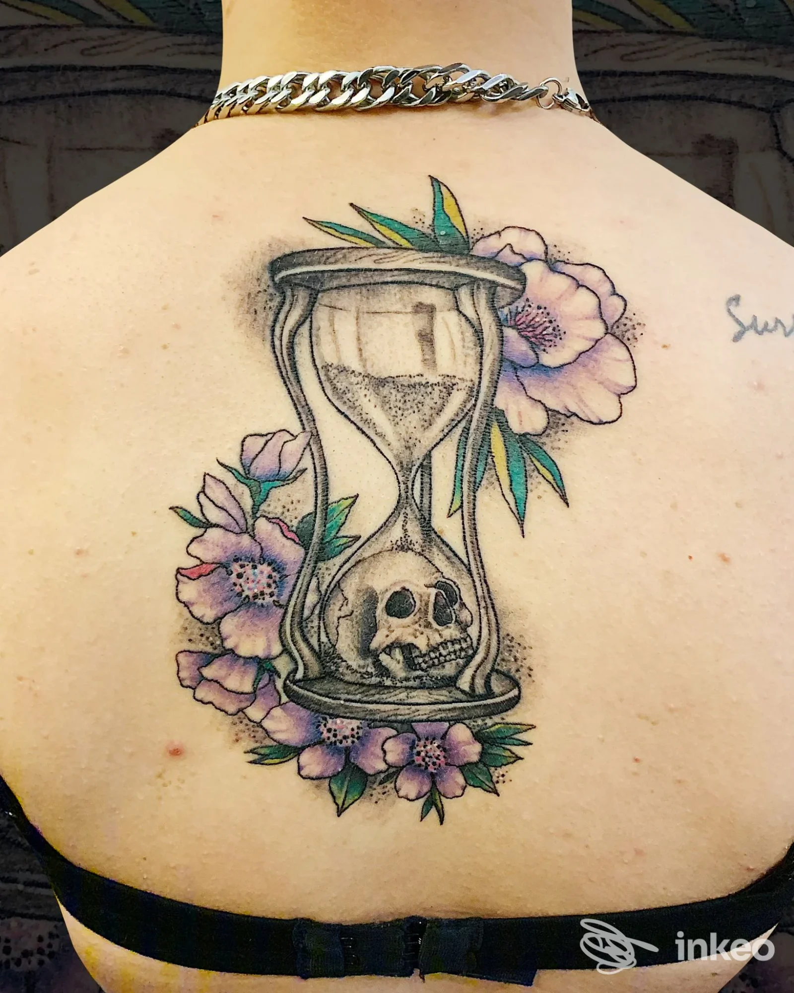 Tatuaggio sul dorso superiore con clessidra e teschio circondato da fiori viola e foglie verdi, visibile direttamente sulla pelle.