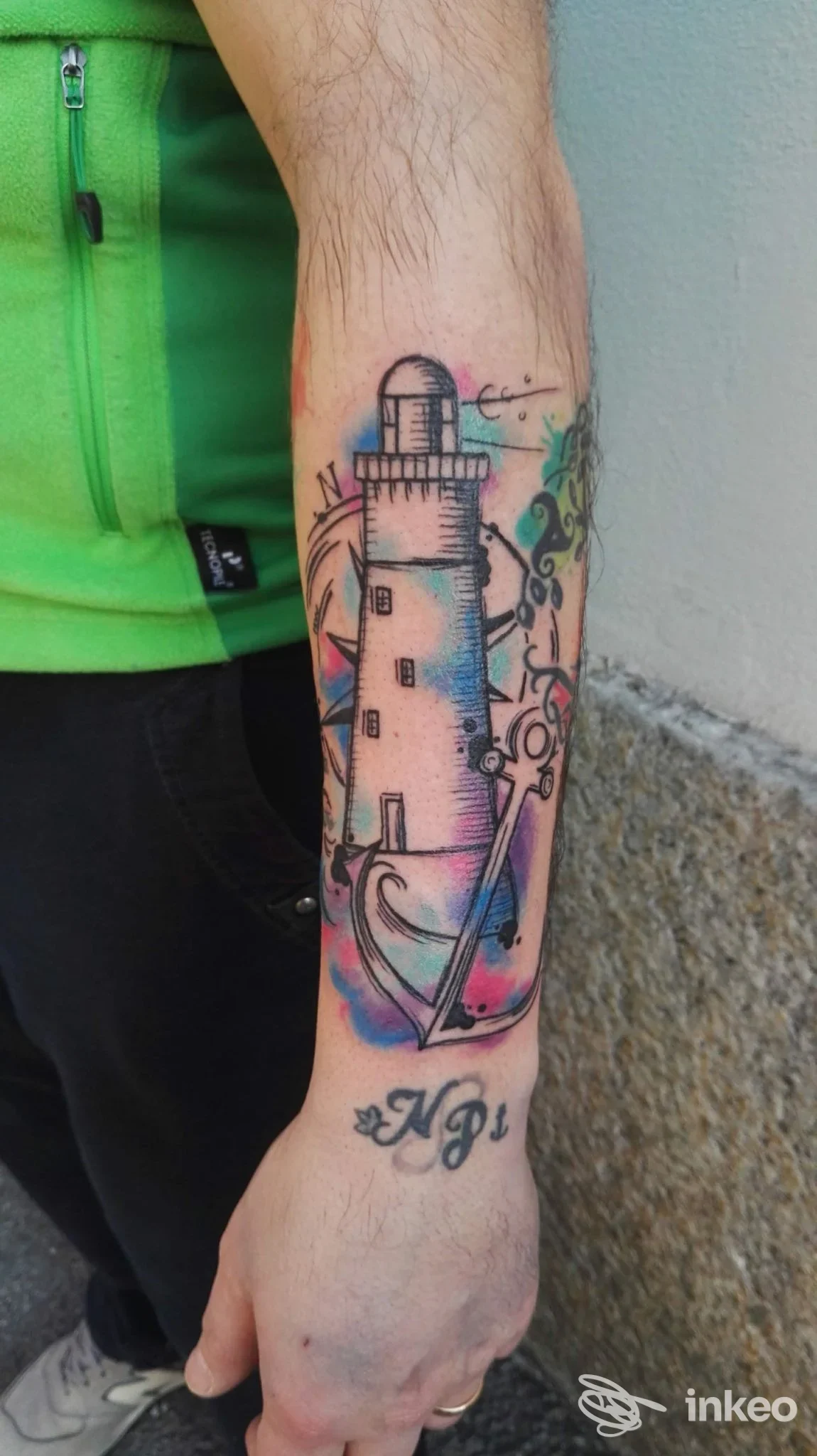 Tattoo a colori di un faro stilizzato sulla parte interna dell'avambraccio, con sfondo acquarellato e dettagli, realizzato su pelle.