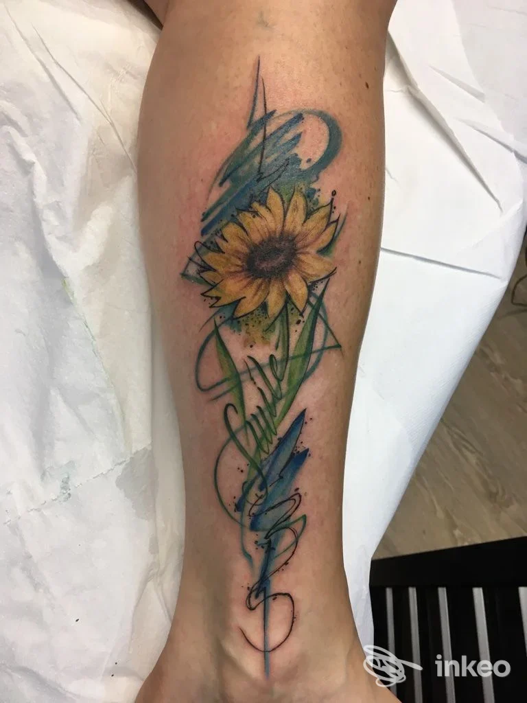 Tatuaggio su avambraccio sinistro raffigurante un fiore di girasole dorato con decorazioni fluide azzurre e verdi lungo la pelle.