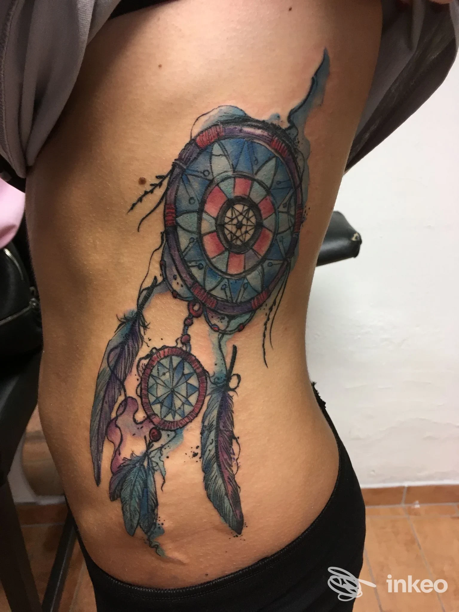 Tattoo visibile sul fianco sinistro raffigurante un dreamcatcher con piume azzurre e dettagli colorati, realizzato direttamente sulla pelle.