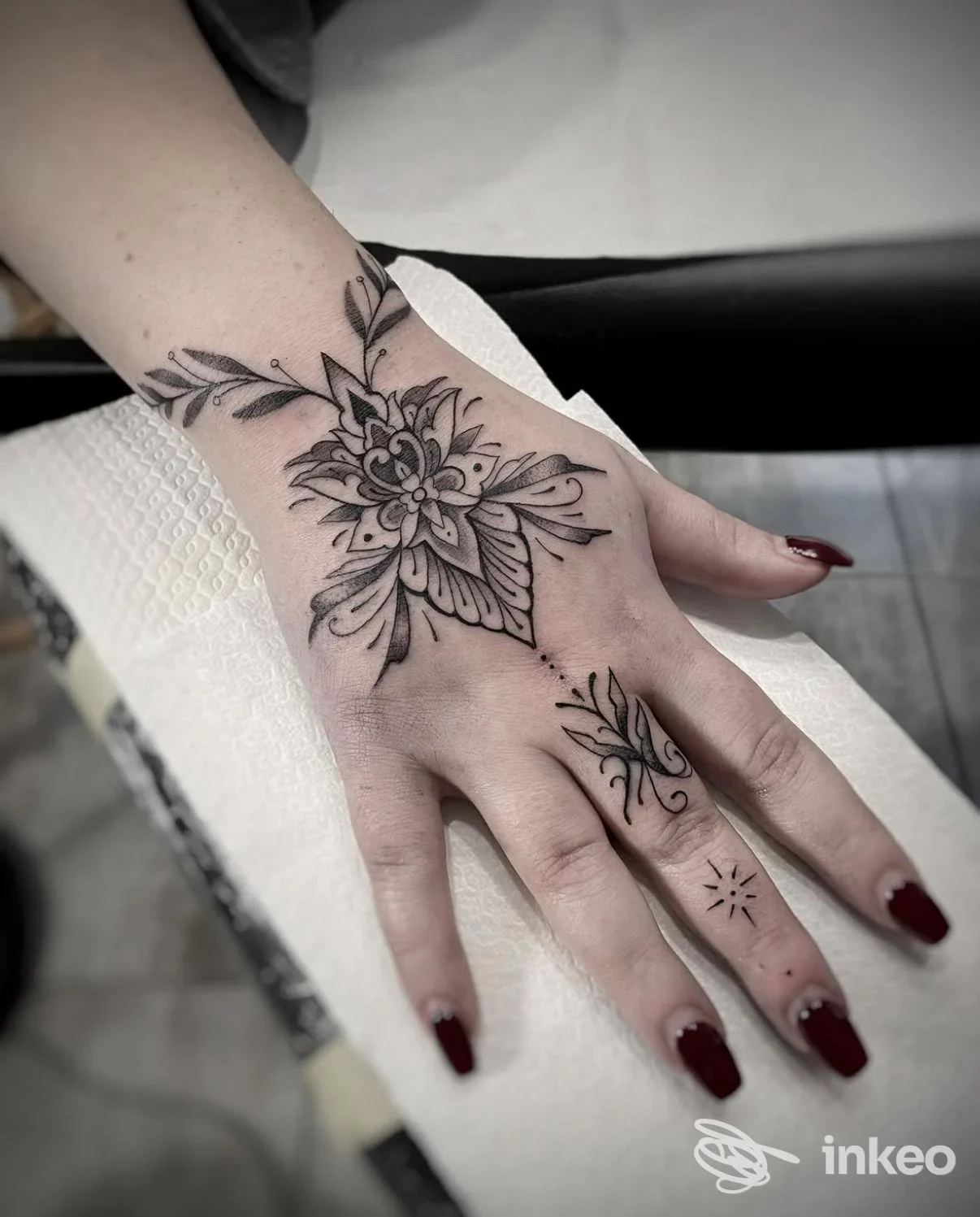 Tattoo sul dorso della mano con motivo floreale dettagliato, eseguito su pelle reale. Unghie verniciate di rosso scuro.