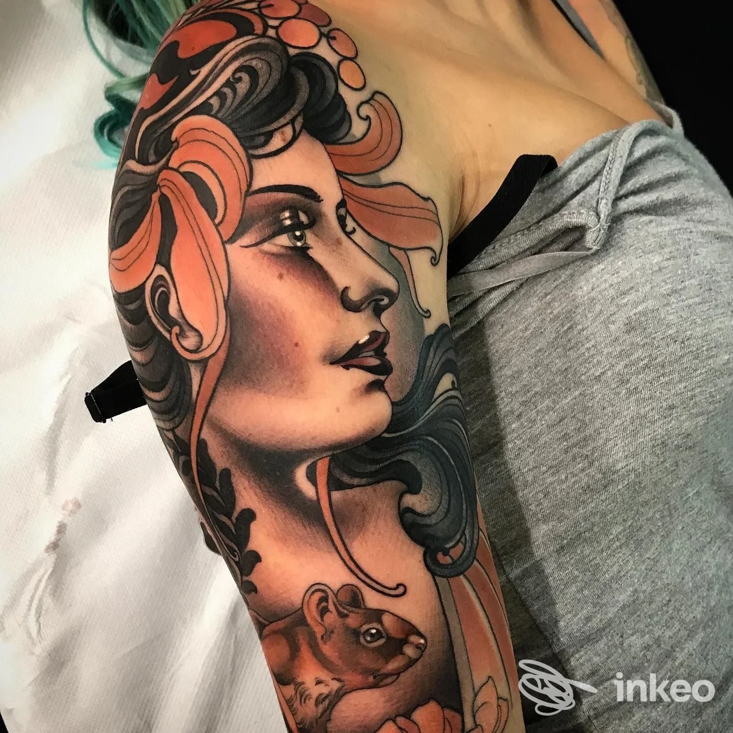 Tatuaggio a braccio raffigurante un profilo femminile stilizzato con capelli ondulati e colori arancioni e neri; in primo piano è presente un piccolo roditore. La scena è un braccio umano reale con la texture della pelle visibile.