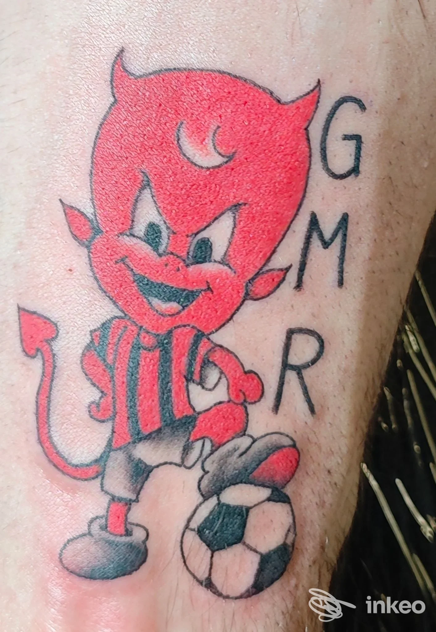 Tatuaggio su gamba raffigurante un diavolo rosso che gioca con un pallone, accompagnato da lettere nere lungo il fianco.