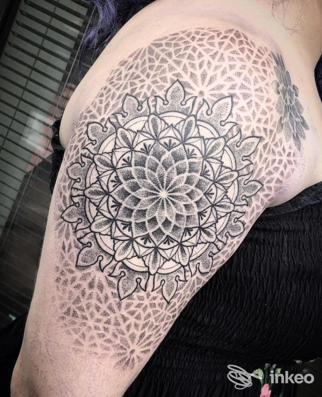 Disegno di mandala geometrico nero, con triangoli e motivi concentrici sull’avambraccio/spalla di una persona, in primo piano.
