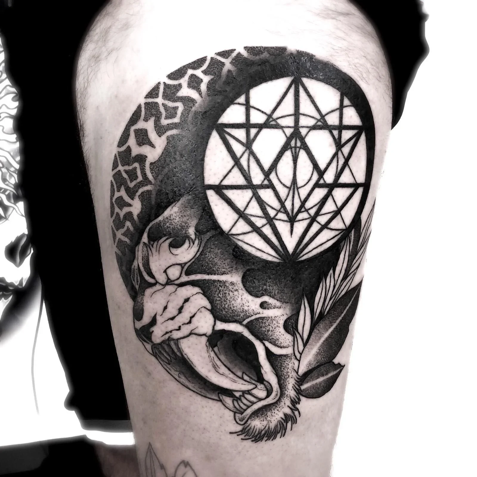 Tattoo geometrico a linee nere su braccio, con cerchio centrale e motivi geometrici, accompagnato da elementi naturali stilizzati.