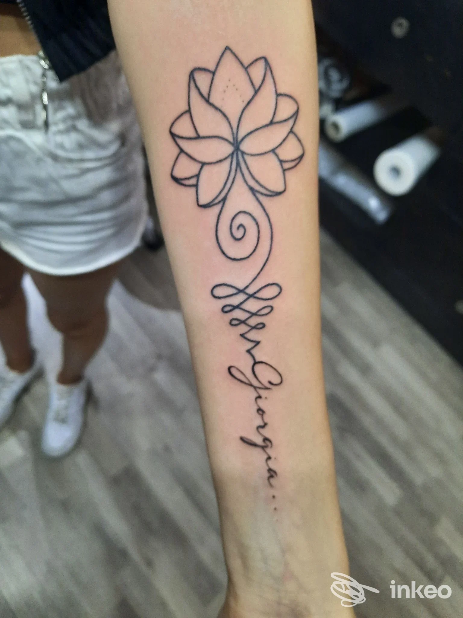 Tatuaggio sull'avambraccio: fiore di loto stilizzato in alto e scritta corsiva lungo la pelle.
