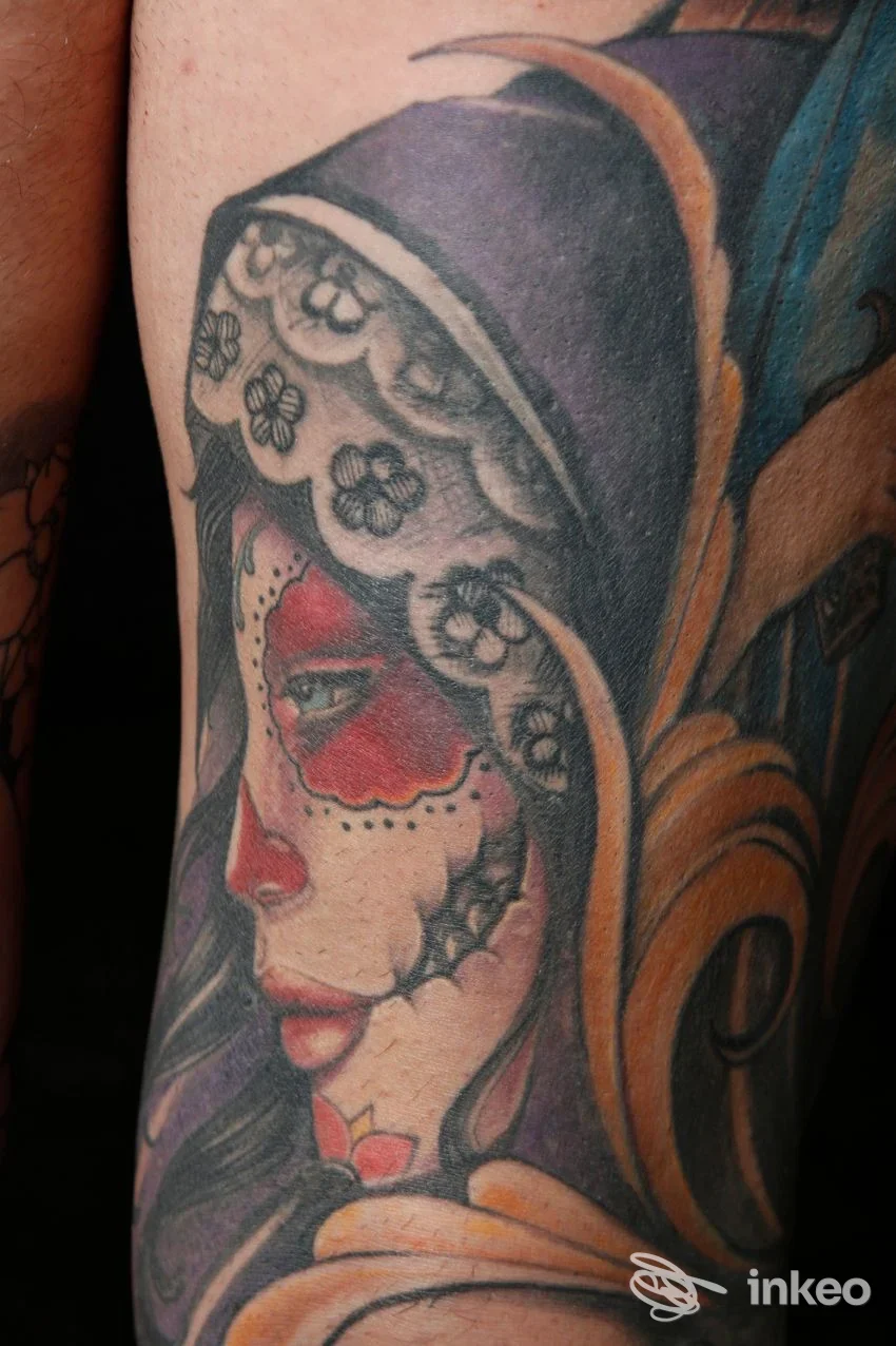 Tattoo a colori in stile Día de los Muertos su braccio, raffigurante una donna con corone di fiori e motivi decorativi, eseguito direttamente sulla pelle.