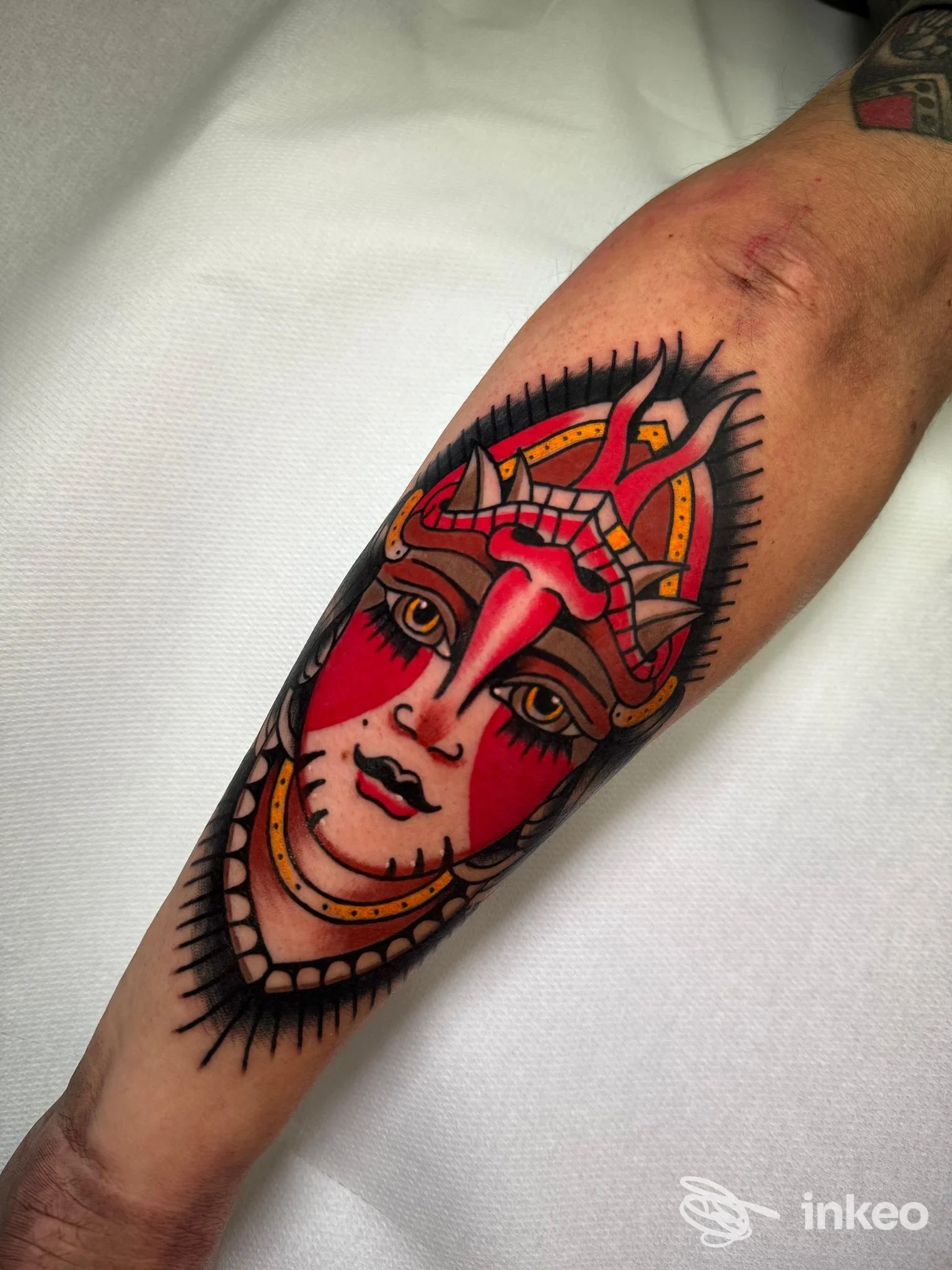 Tatuaggio su avambraccio raffigurante un volto femminile stilizzato con colori rossi e contorni neri, con ornamenti dorati, realizzato sulla pelle umana in studio.