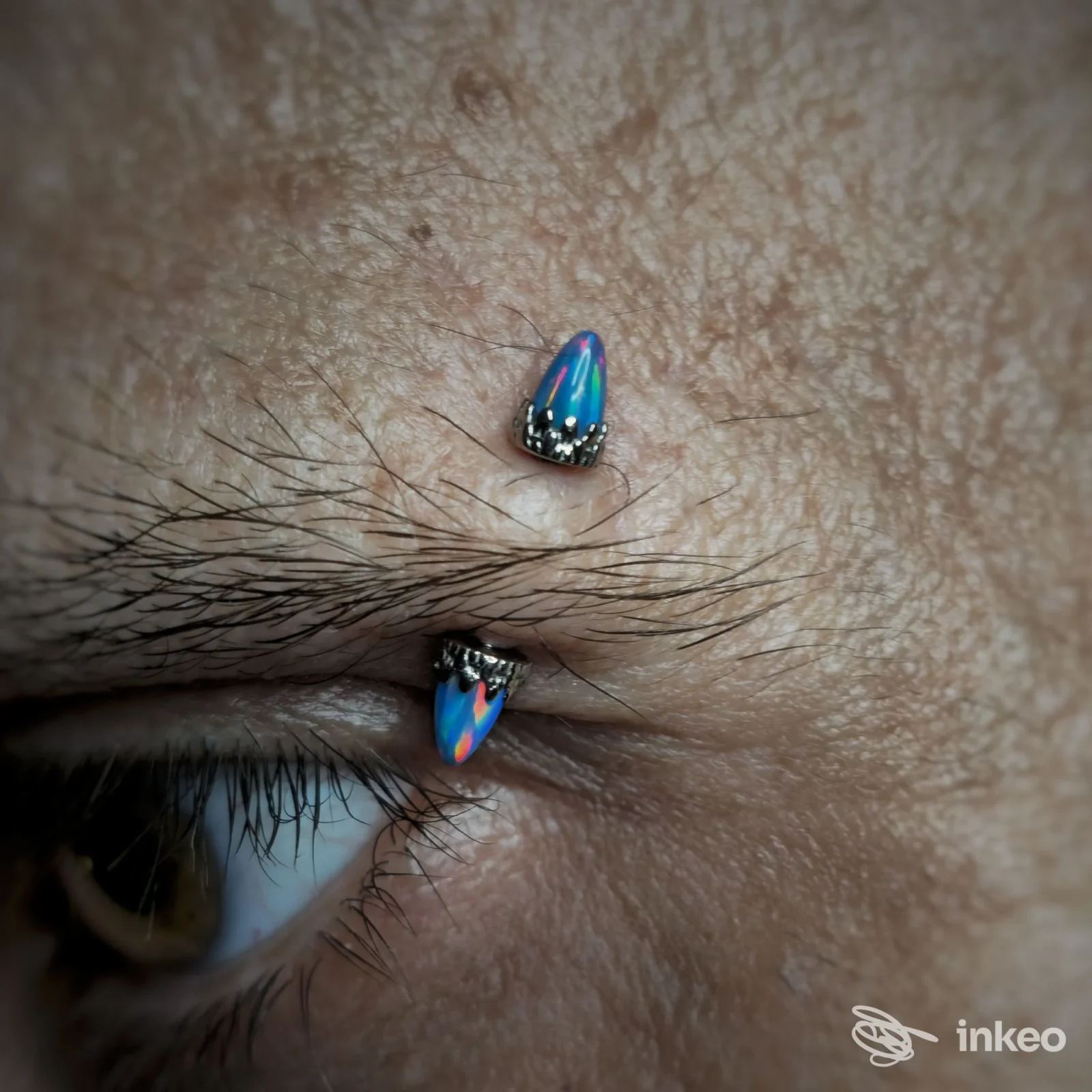 Primo piano su due piercing colorati posizionati vicino all'occhio su pelle visibile; i gioielli hanno base metallica con teste a forma di goccia blu.