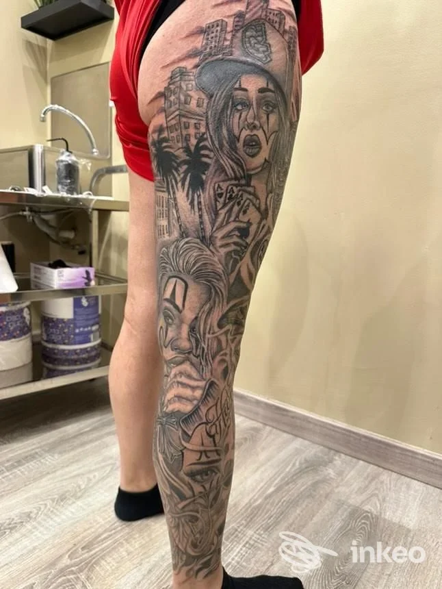 Tattooing permanente sulla gamba di una persona: un braccio lungo completo con dettagli in bianco e nero che mostrano personaggi, edifici e paesaggi stilizzati, eseguito su pelle reale; l’angolo ritrae la gamba sinistra con sfondo interno di studio.