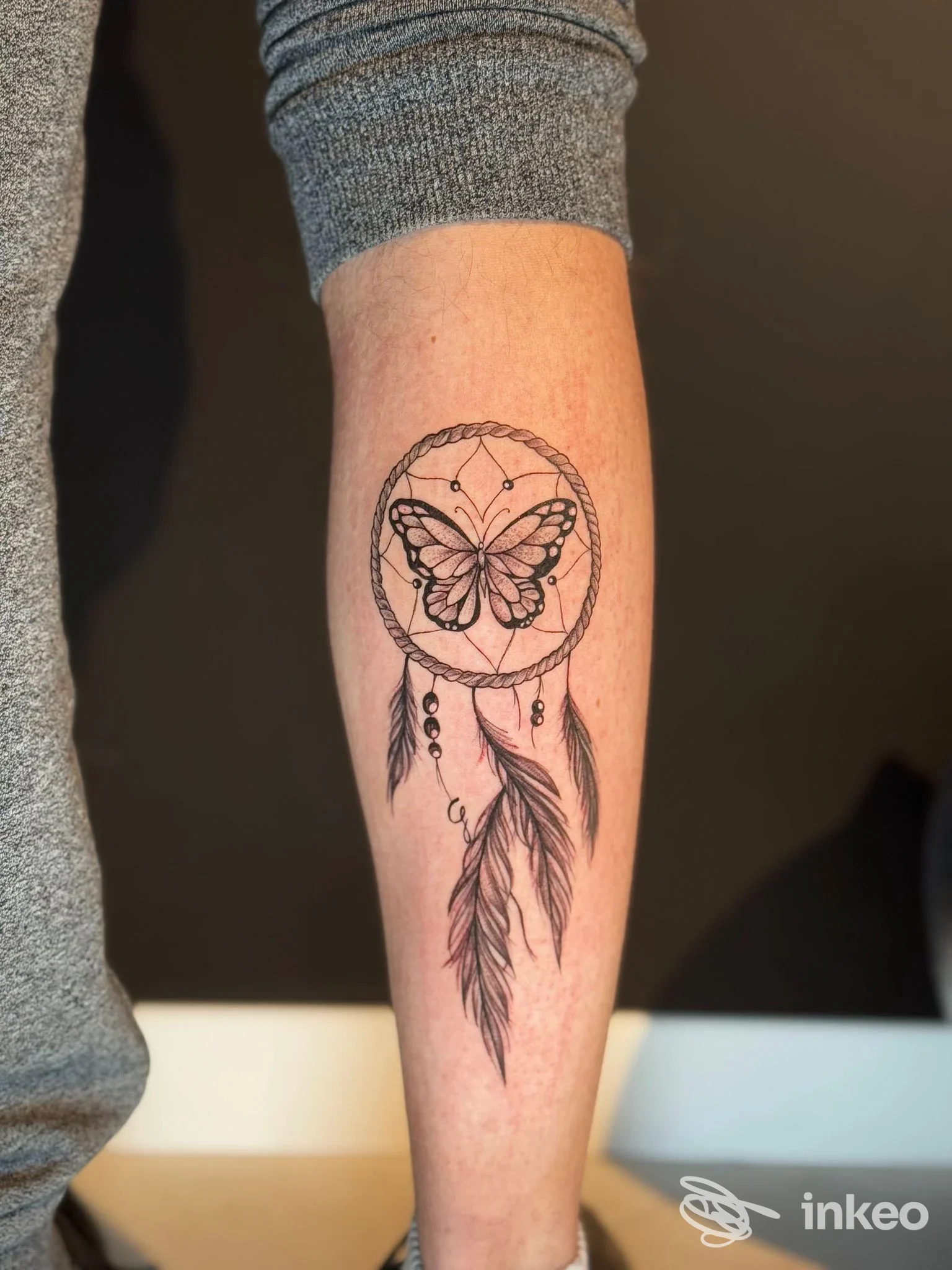 Tattoo reale sul braccio raffigurante una farfalla inserita in un cerchio stile dreamcatcher, con piume pendenti.