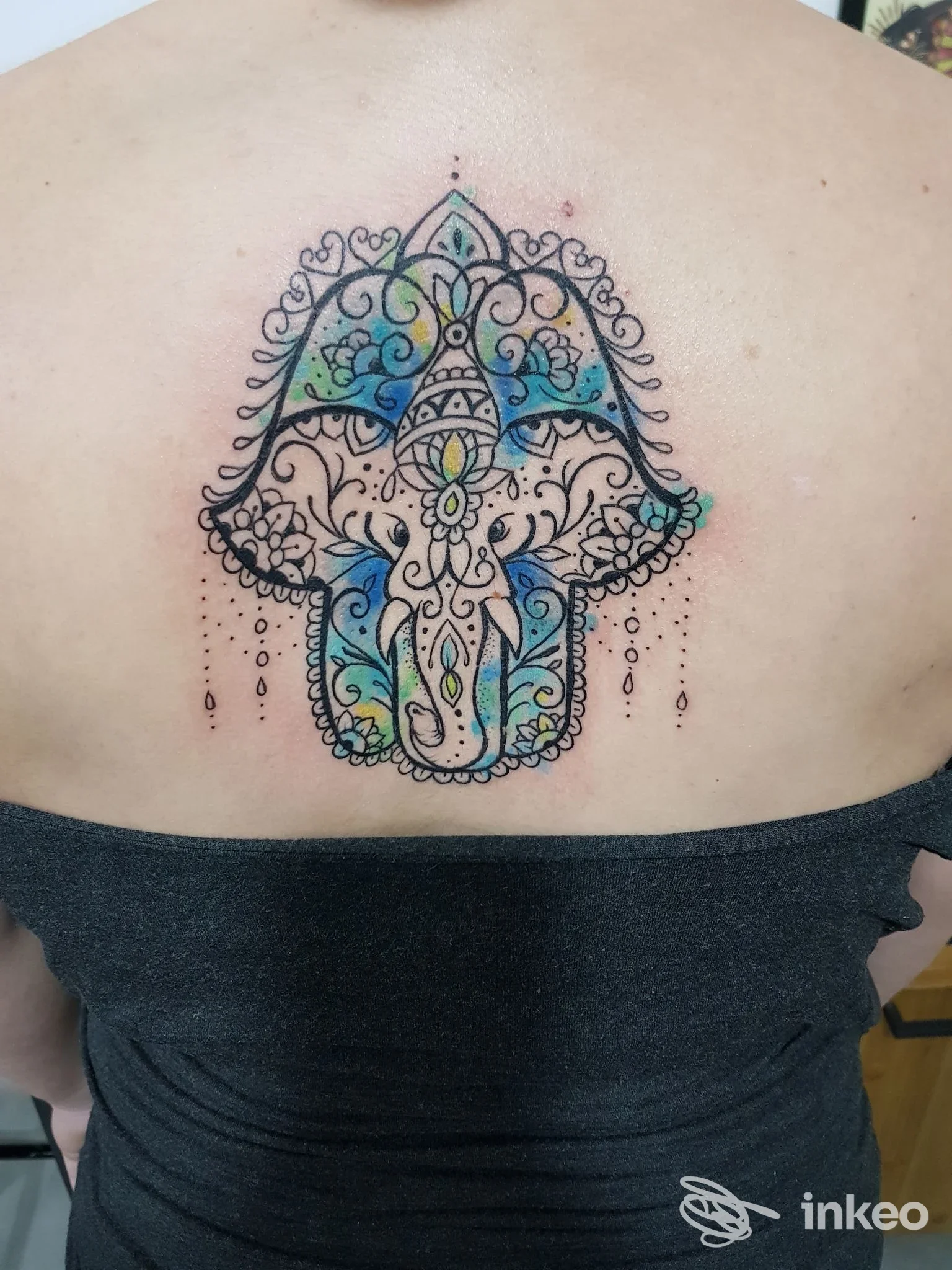 Tattoo colorato sul dorso di una persona, design ornato con testa di elefante in stile mandala, contorni neri e riempimenti azzurri, con pendenti decorativi lungo la parte inferiore.