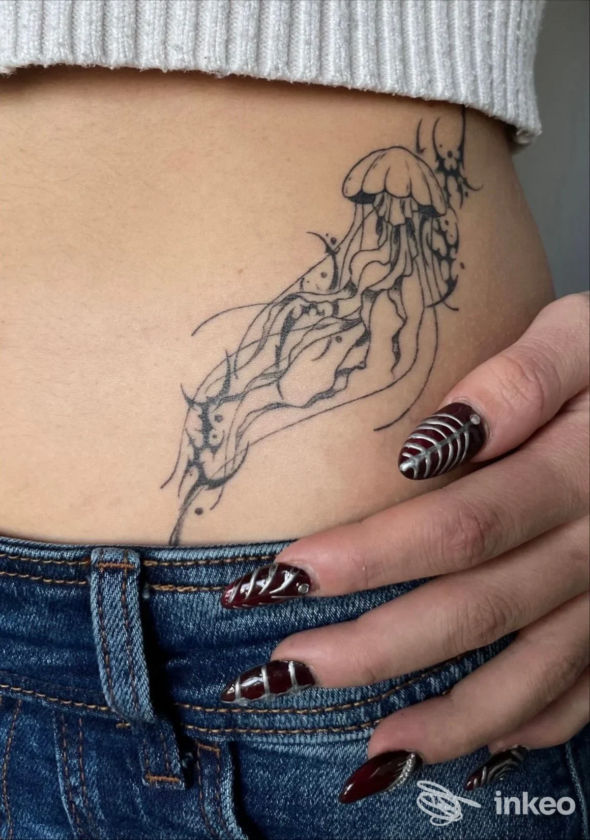 Tatuaggio nero su pelle chiara lungo il fianco, raffigurante una medusa stilizzata con linee fluide, realizzato sulla pelle reale. prima piano mostra mano con unghie decorate e jeans.