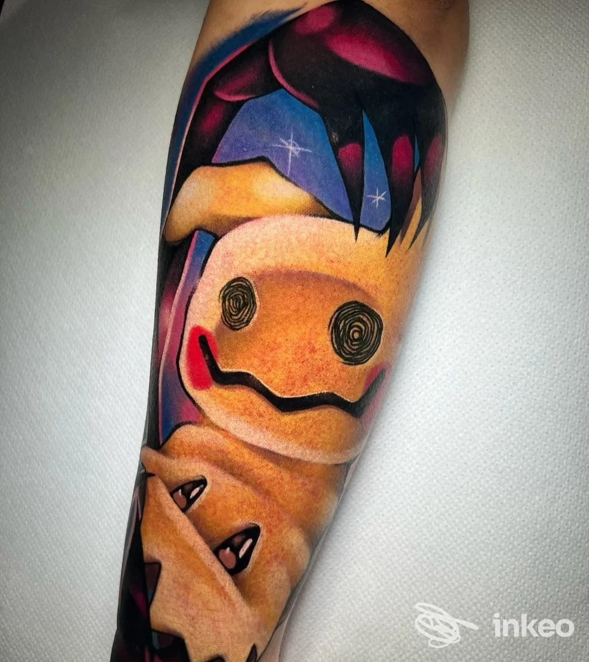 Tatuaggio a colori vividi su avambraccio: personaggio cartoonesco stilizzato con volto semplificato, linee nette e sfumature; sfondo chiaro, colori arancioni, viola e nere.