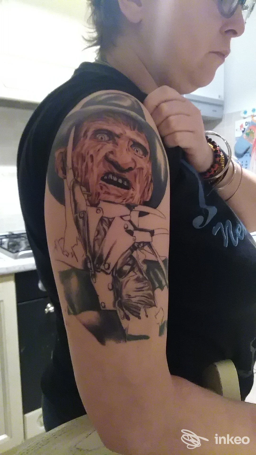 Tatuaggio realistico in bianco e nero su braccio superiore, raffigurante una figura umana dall’espressione intensa; visibile spalla e braccio, individuo in abiti casual.