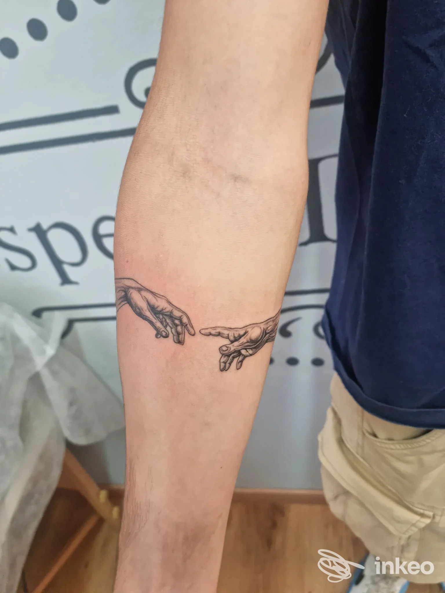 Dettaglio di un avambraccio con tatuaggio raffigurante due mani che si sfiorano; la pelle è visibile e non compaiono volti interi o elementi di social network.