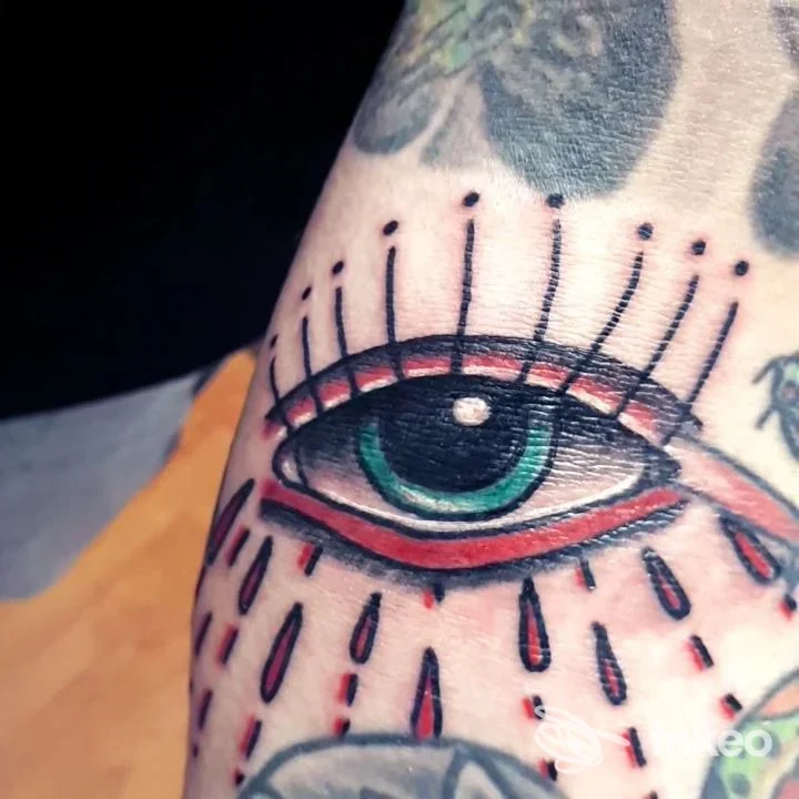 Tattoo sull'avambraccio raffigurante un occhio stilizzato con iris verde e lacrime rosse, stile grafico.
