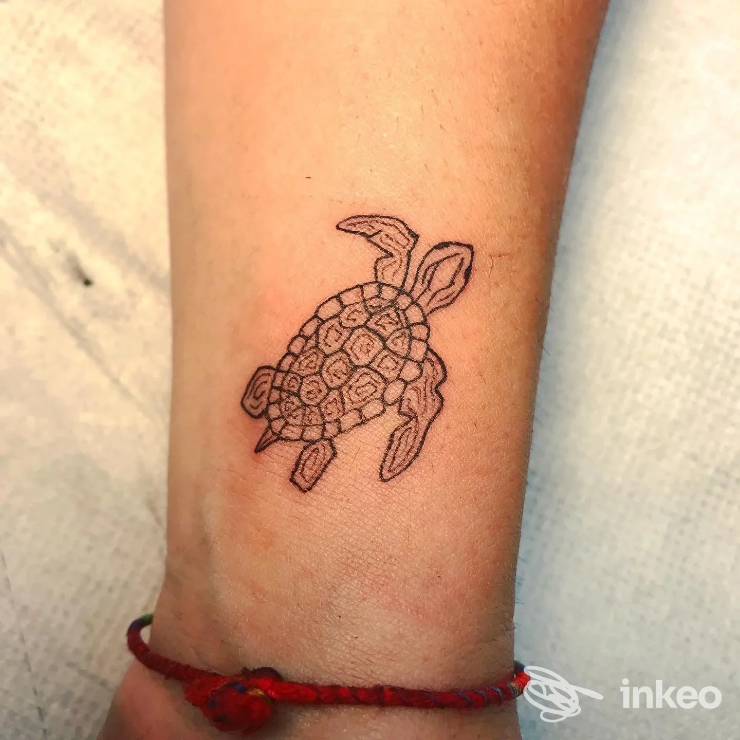 Tattoo lineare di una tartaruga su avambraccio, contorni neri e riempimento minimo, sfondo chiaro e braccialetto rosso visibile.