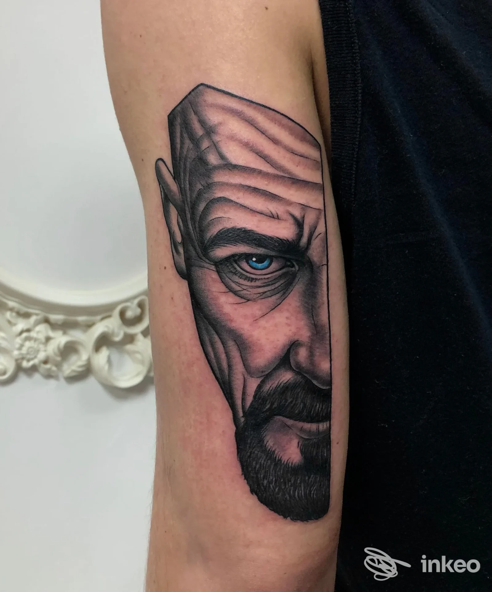 Tatuaggio realistico su pelle: ritratto parziale di un uomo con barba e occhio azzurro sull'avambraccio.