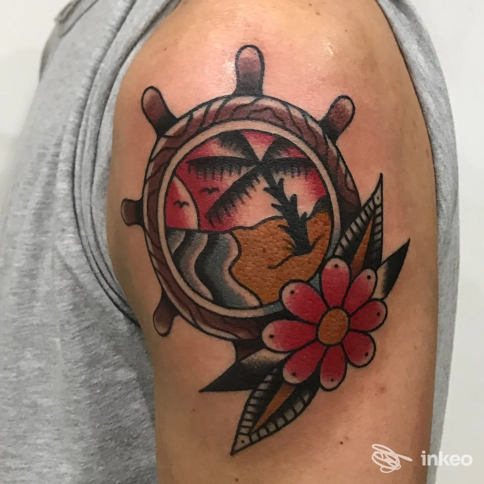 Tattoo sull'avambraccio raffigurante una scena marina tropicale entro una ruota, con palma e tramonto, accompagnata da un fiore rosa. realizzato sulla pelle.
