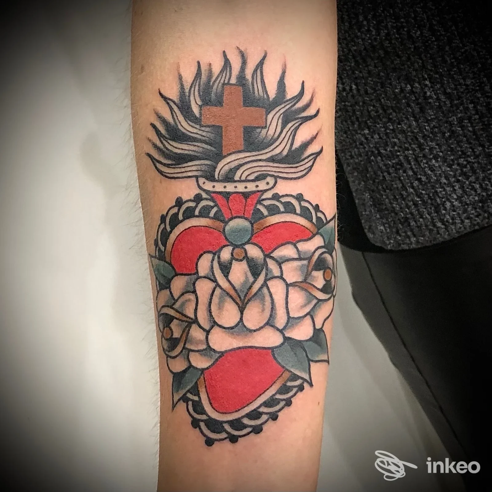 Tattoo sull'avambraccio raffigurante una croce con fiamme, cuore e rose rosse, contorni neri netti, stile tradizionale; la pelle è visibile senza elementi di influencer o testo.