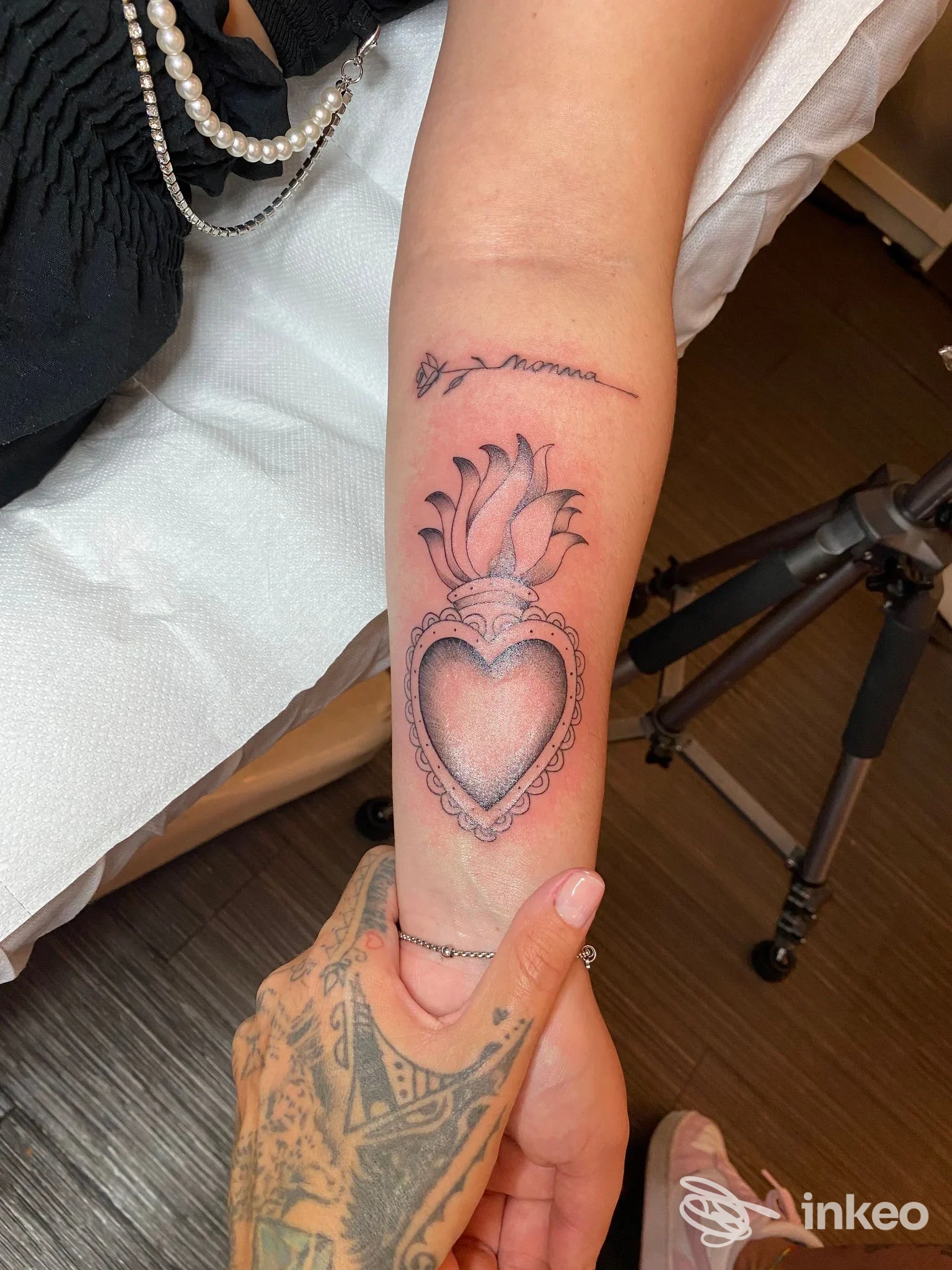 Braccio inferiore di una persona con un tatuaggio a cuore decorato sull’avambraccio, con una scritta sottile in corsivo sopra, senza viso visibile.