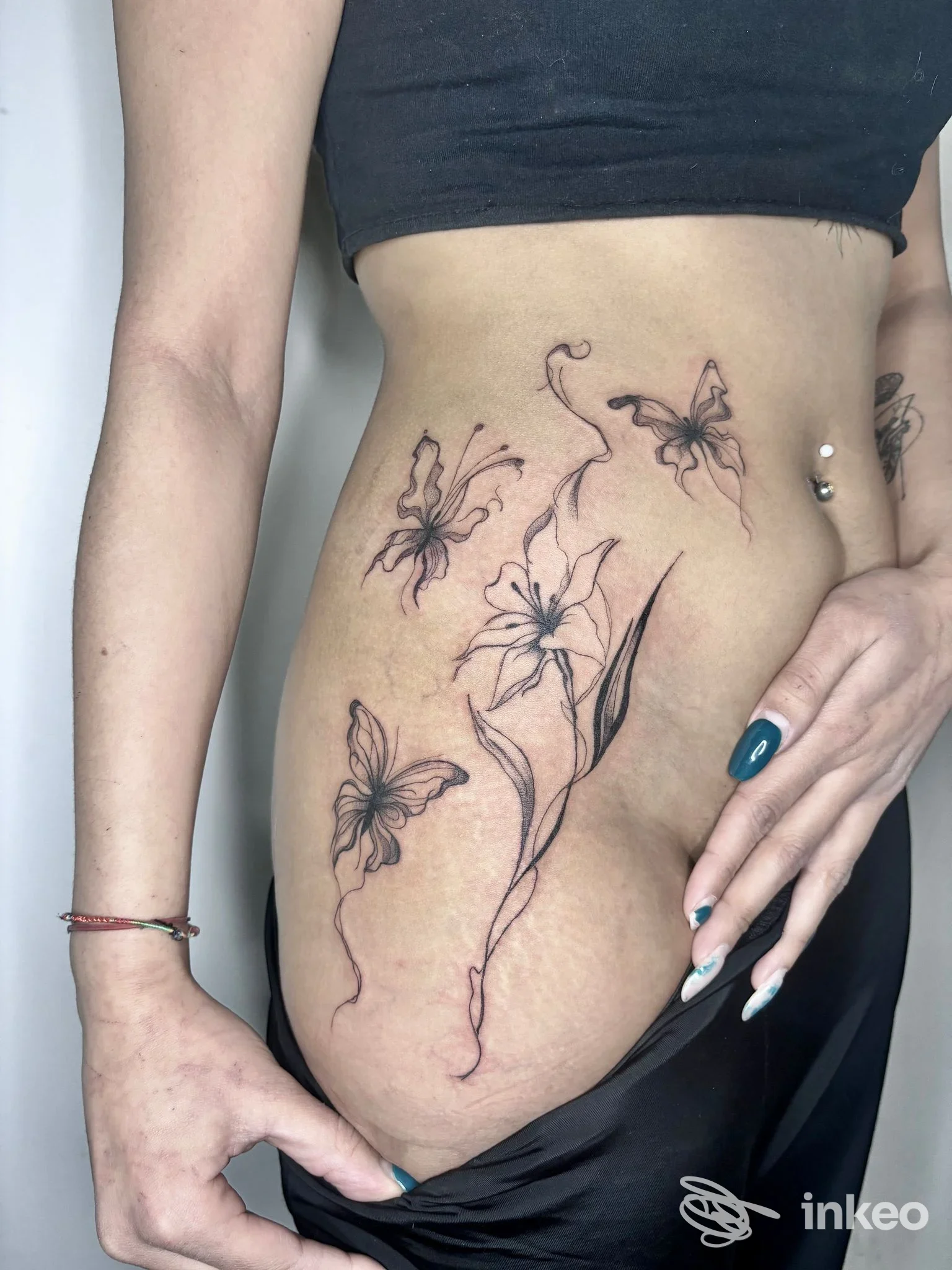 Tattoo realistico su addome/fianco sinistro: fiori stilizzati e farfalle in nero, visibile su pelle reale di una persona.