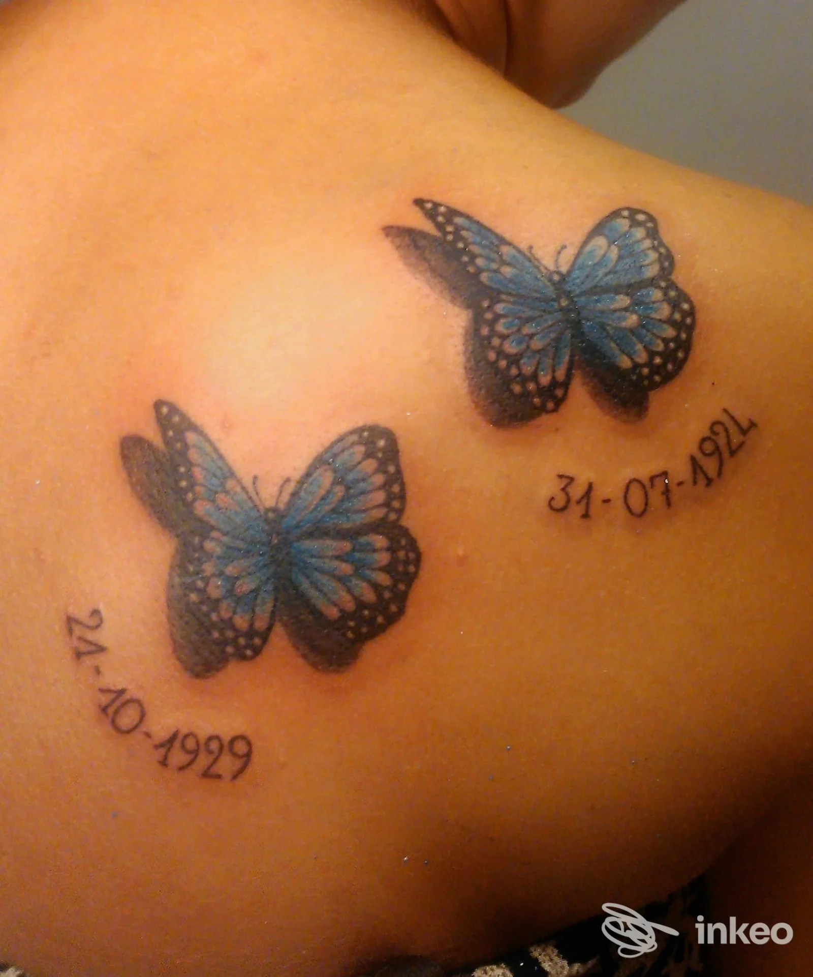 Due farfalle blu tatuate sulla spalla, con date curve accanto, realizzate sulla pelle.