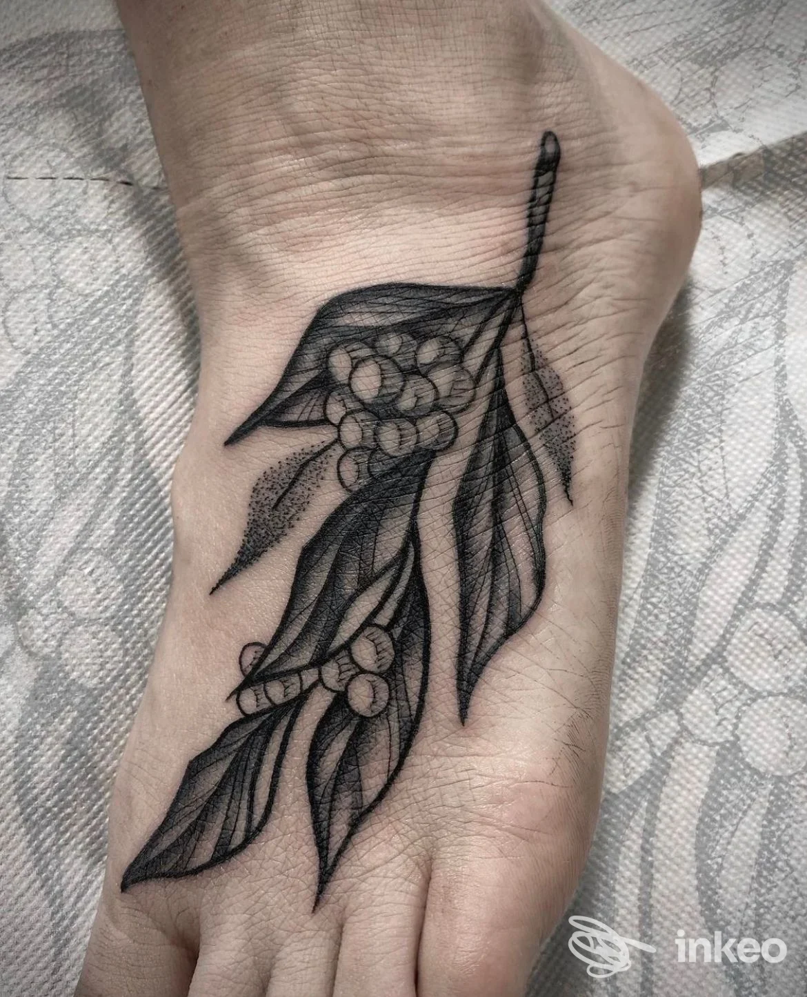 Tattoo nera su polso/mano raffigurante un ramo con foglie e fiori, realizzata sulla pelle; visibile pezzo di tessuto sullo sfondo.