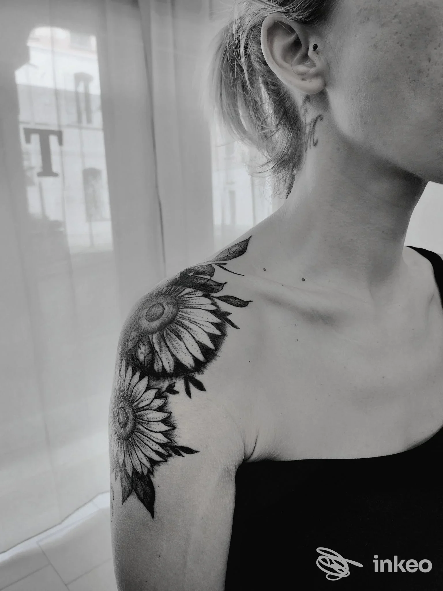 Tattoo reale visibile su spalla sinistra: motivo di girasole in bianco e nero, con dettagli di petali lungo la spalla; parte del volto e orecchio visibili.