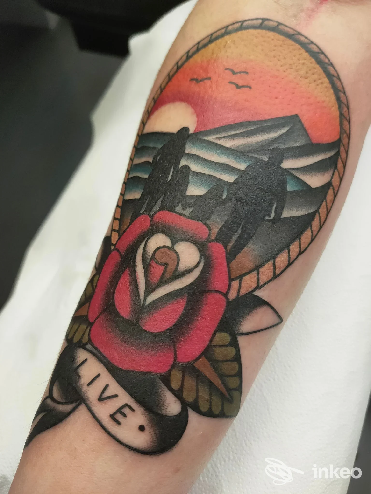 Tatuaggio sul braccio raffigurante una rosa rossa con sfondo paesaggistico al tramonto; colori vividi e contorni netti su pelle reale, nessun testo visibile.