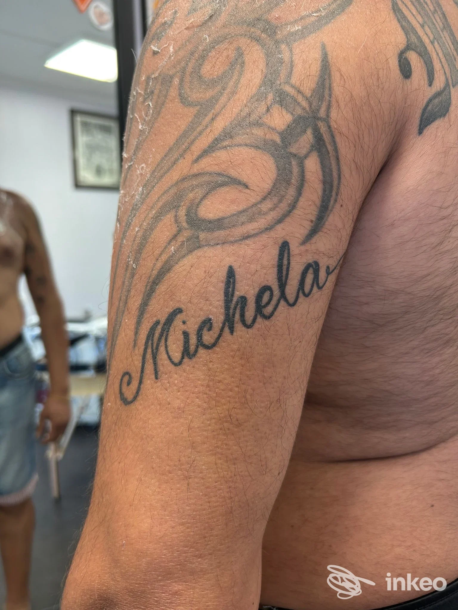 Braccio sinistro con tatuaggio scriptato 'Michela' in corsivo e motivi decorativi, visibile sullo sfondo di uno studio, senza volto completo.