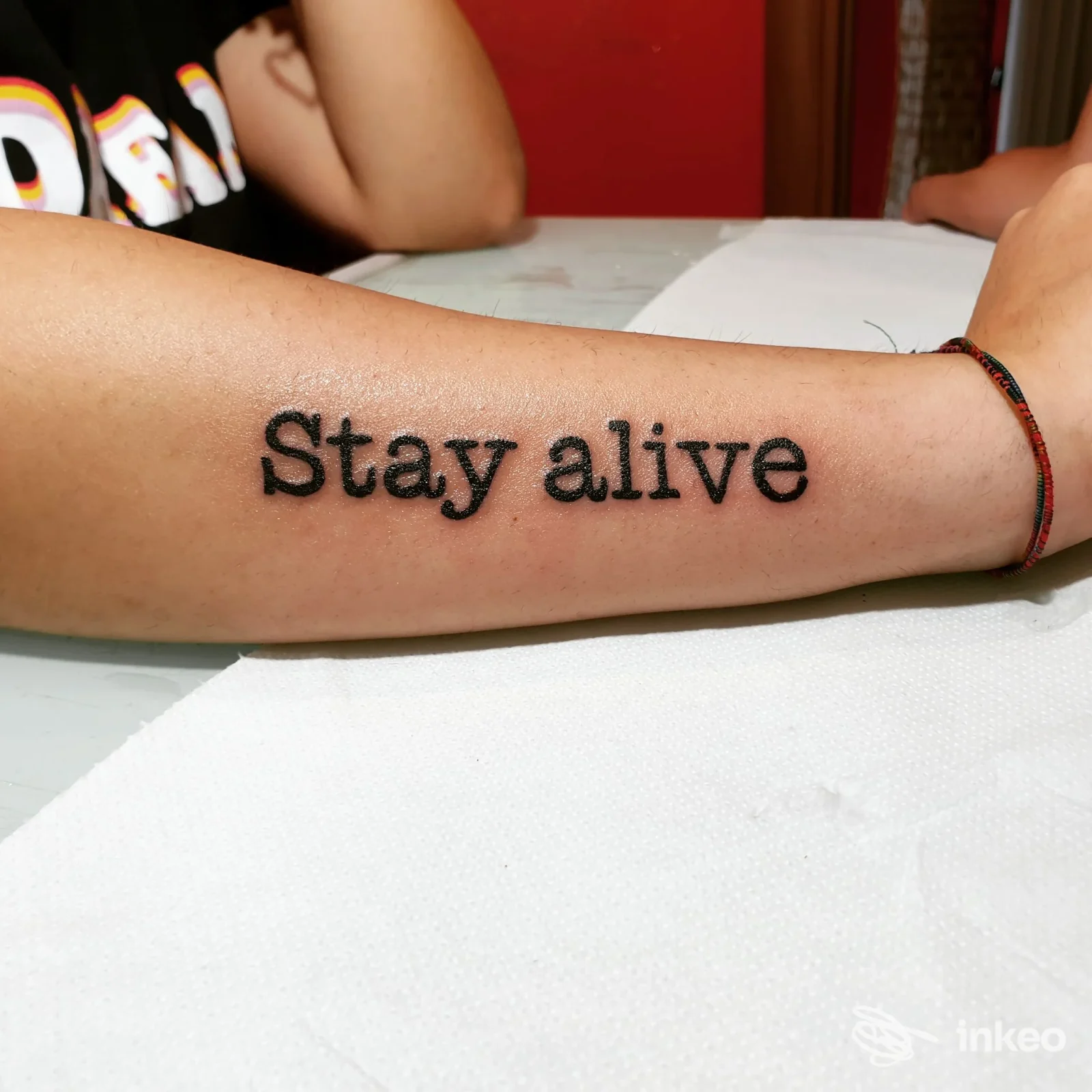 Avambraccio con tatuaggio nero leggibile 'Stay alive' eseguito sulla pelle, visibile durante una sessione in studio Real Tattoo.