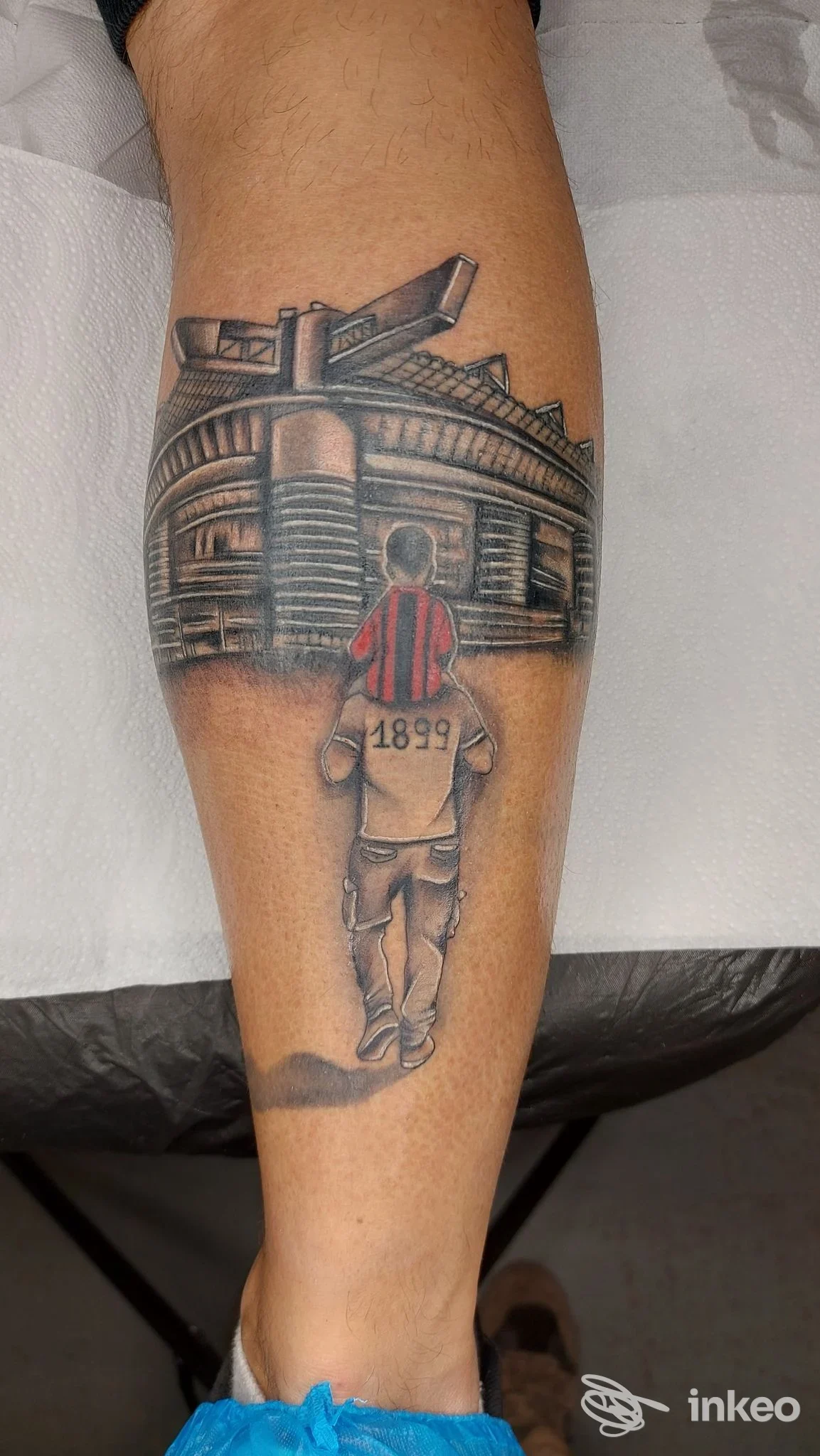 Tatuaggio sul polpaccio raffigurante una persona che cammina verso una struttura simile a uno stadio, con l'anno 1899 stampato sulla schiena, realizzato in tonalità black and gray.