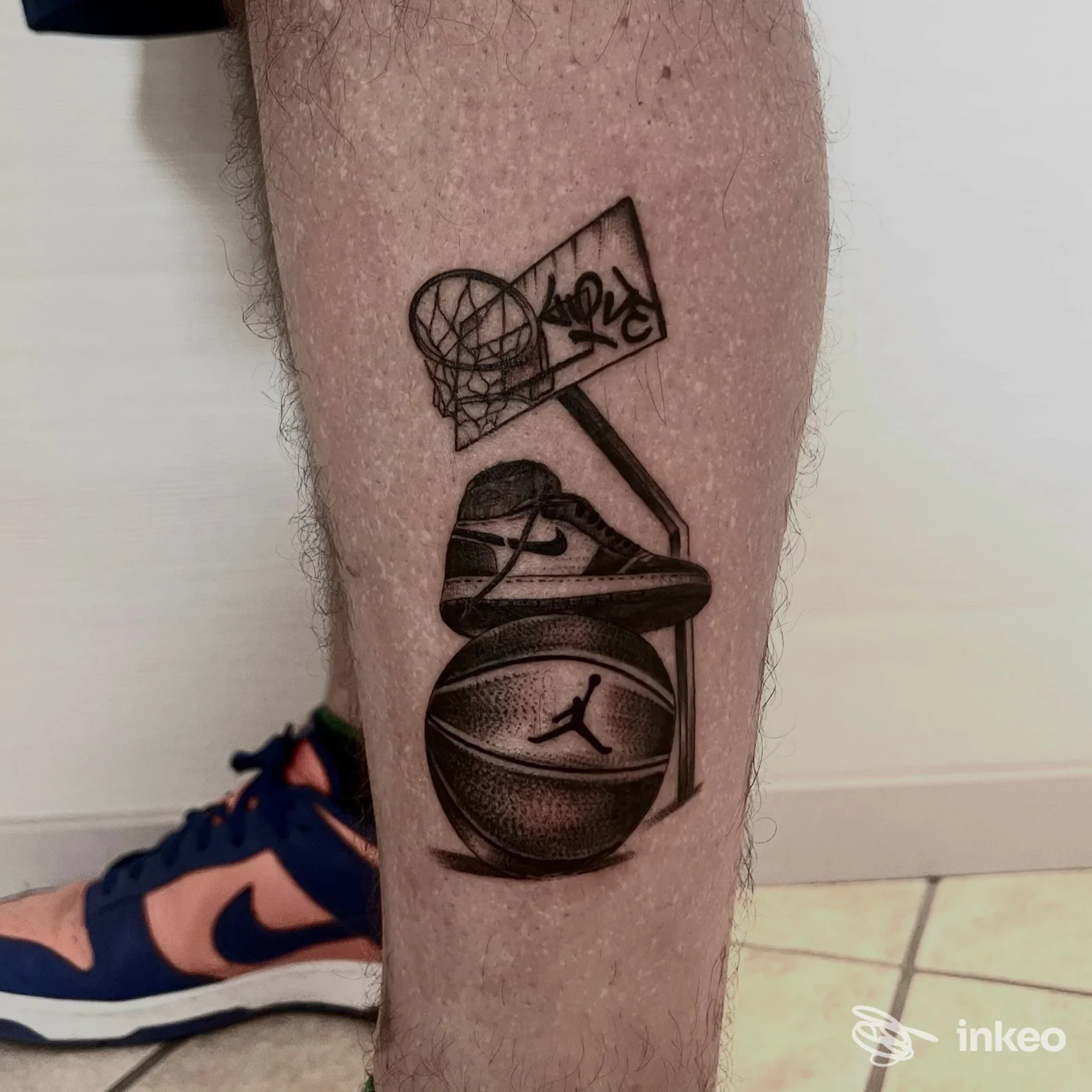 Tattoo su gamba inferiore raffigurante palla da basket dentro canestro e una scarpa appoggiata, in bianco e nero.