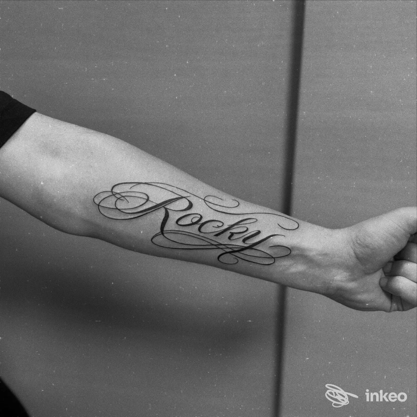 Tatuaggio script sul braccio: nome 'Rocky' scritto in elegante corsivo sull'avambraccio, visibile senza volto, sfondo neutro.