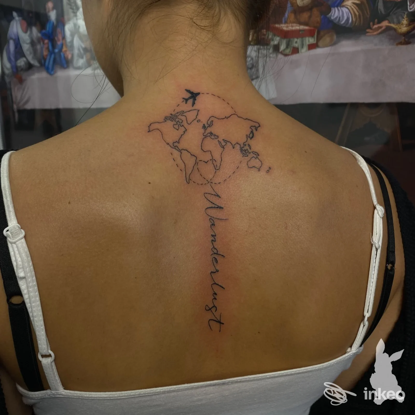 Tattoo sulla schiena: mappa del mondo con un aereo e la parola wanderlust scritta verticalmente lungo la colonna vertebrale.