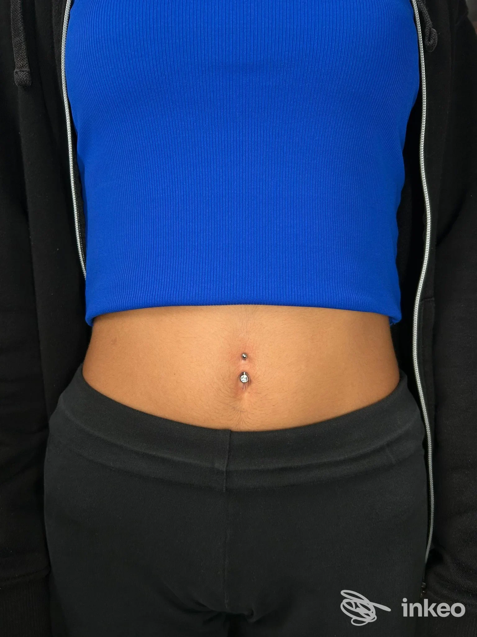 Piercing all'ombelico con due gemme su pelle reale, visibile sull'addome di una persona: cropped blu e giacca nera senza viso.