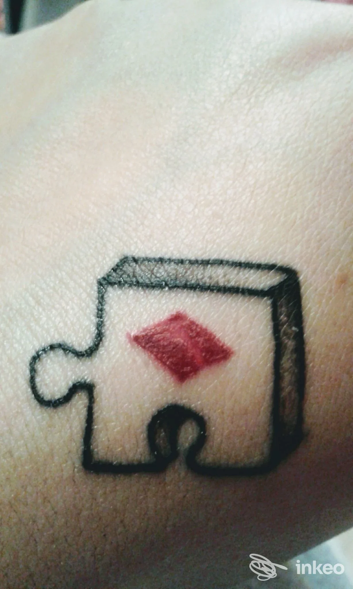 Piccolo tatoo su polso raffigurante un pezzo di puzzle con una gemma rossa, contorno nero, tessitura della pelle visibile.