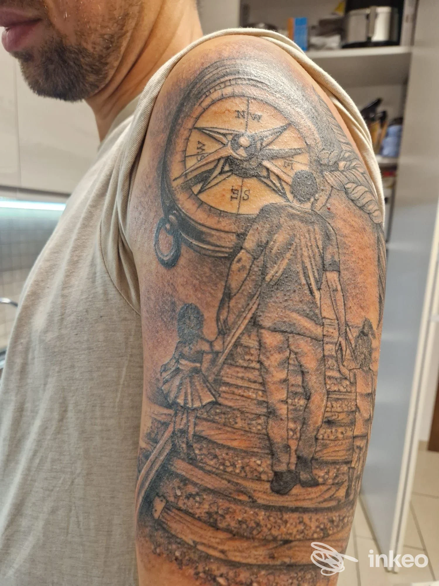 Braccio di un uomo con un grande tatuaggio nero e grigio che mostra una scena con una persona che cammina sui binari e una bussola, in ambientazione di studio tatuaggi.