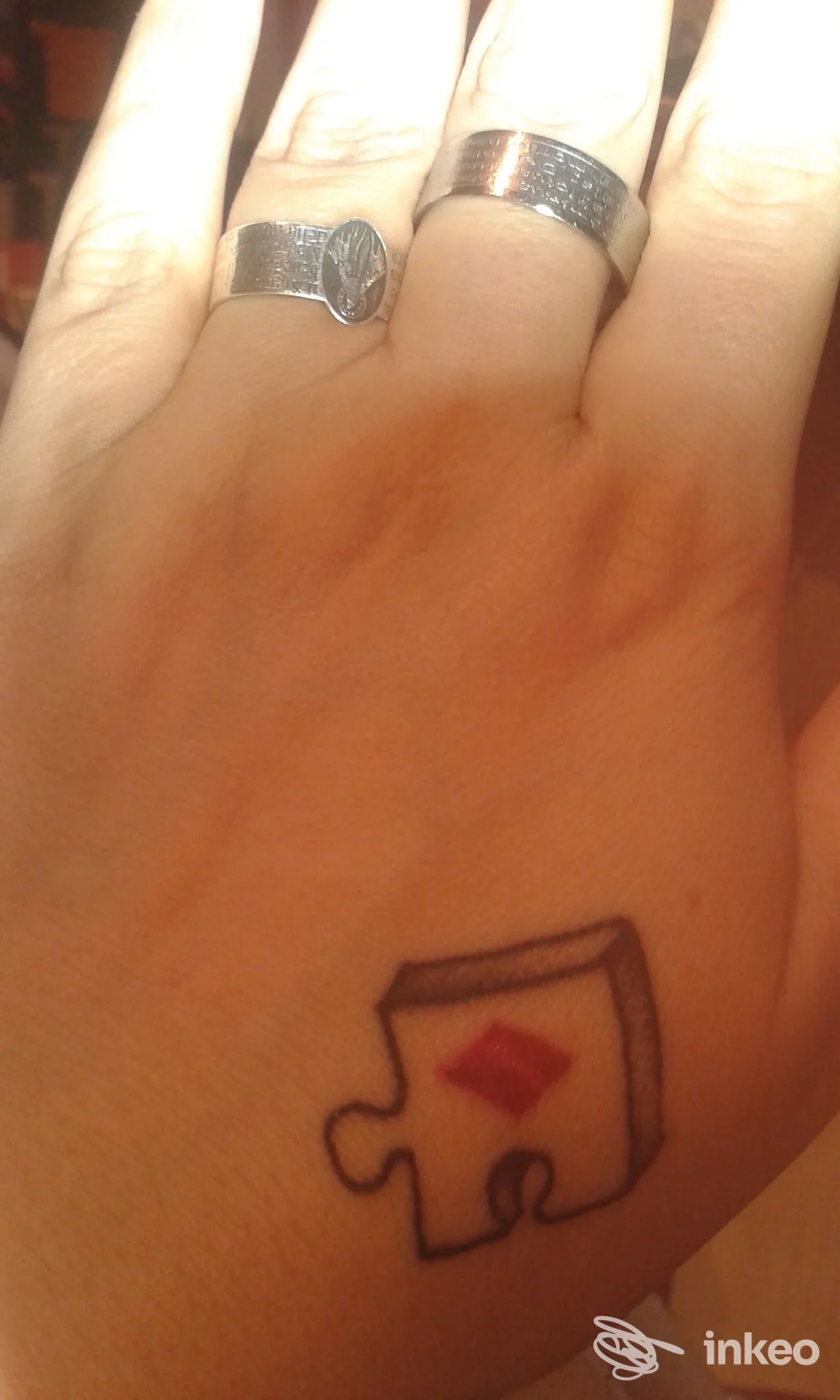 Mano con piccola tatuatura stilizzata a forma di pezzo di puzzle con simbolo rosso sul dorso della mano, indossando anelli, studio Masa Tattoo a Inveruno