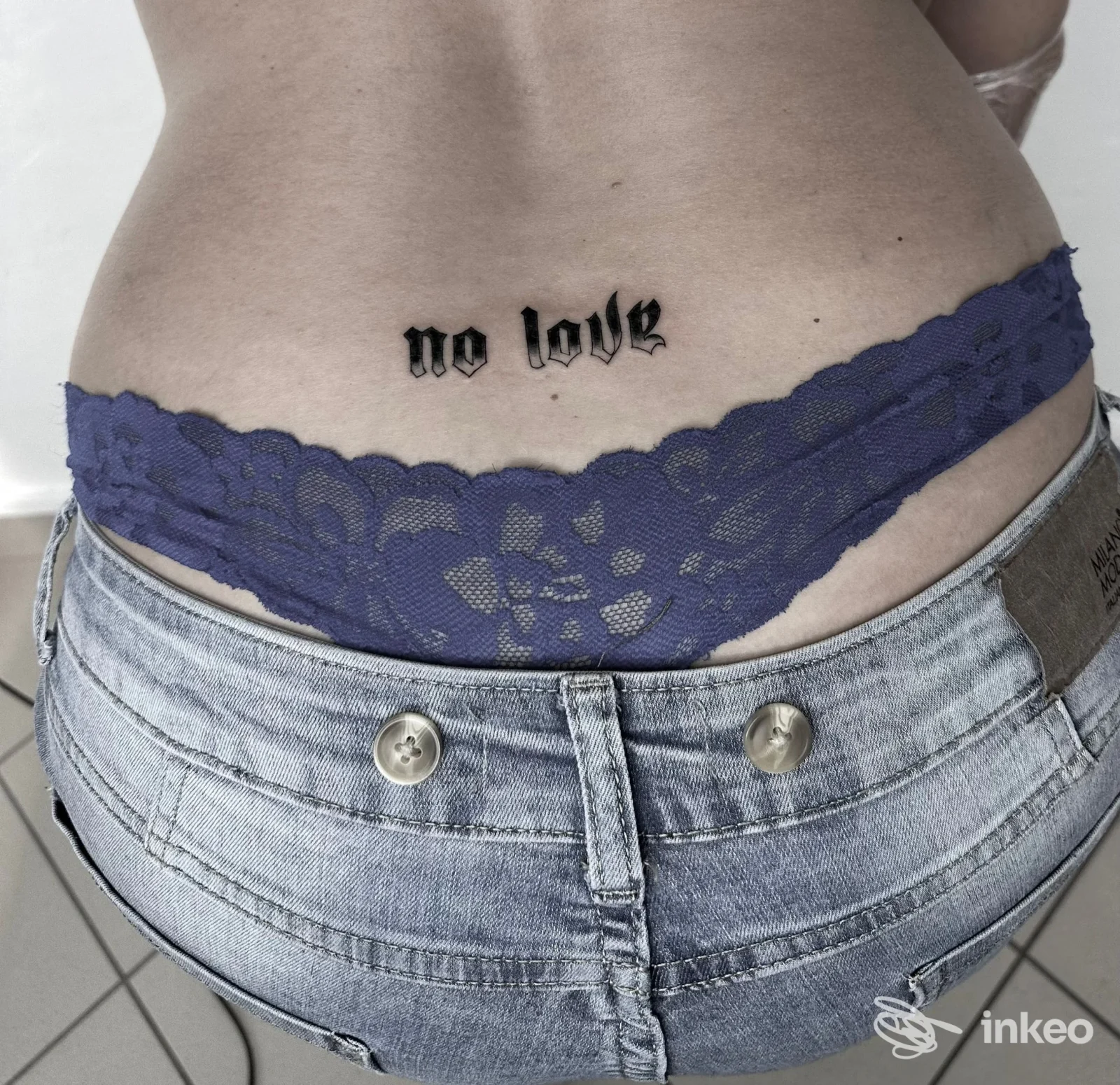 Tattoo sul fianco/lombare con la scritta "no love" in stile gotico, visibile sopra la cintura dei jeans viola.