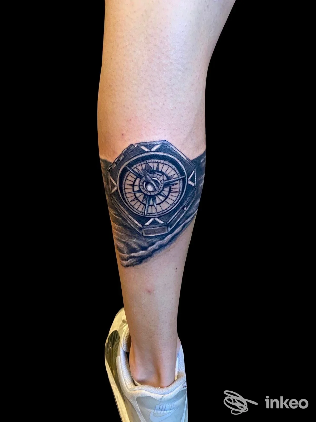 Tattoo visibile sulla gamba di una persona: una bussola/rosa dei venti in bianco e nero posizionata sul polpaccio, sfondo scuro.