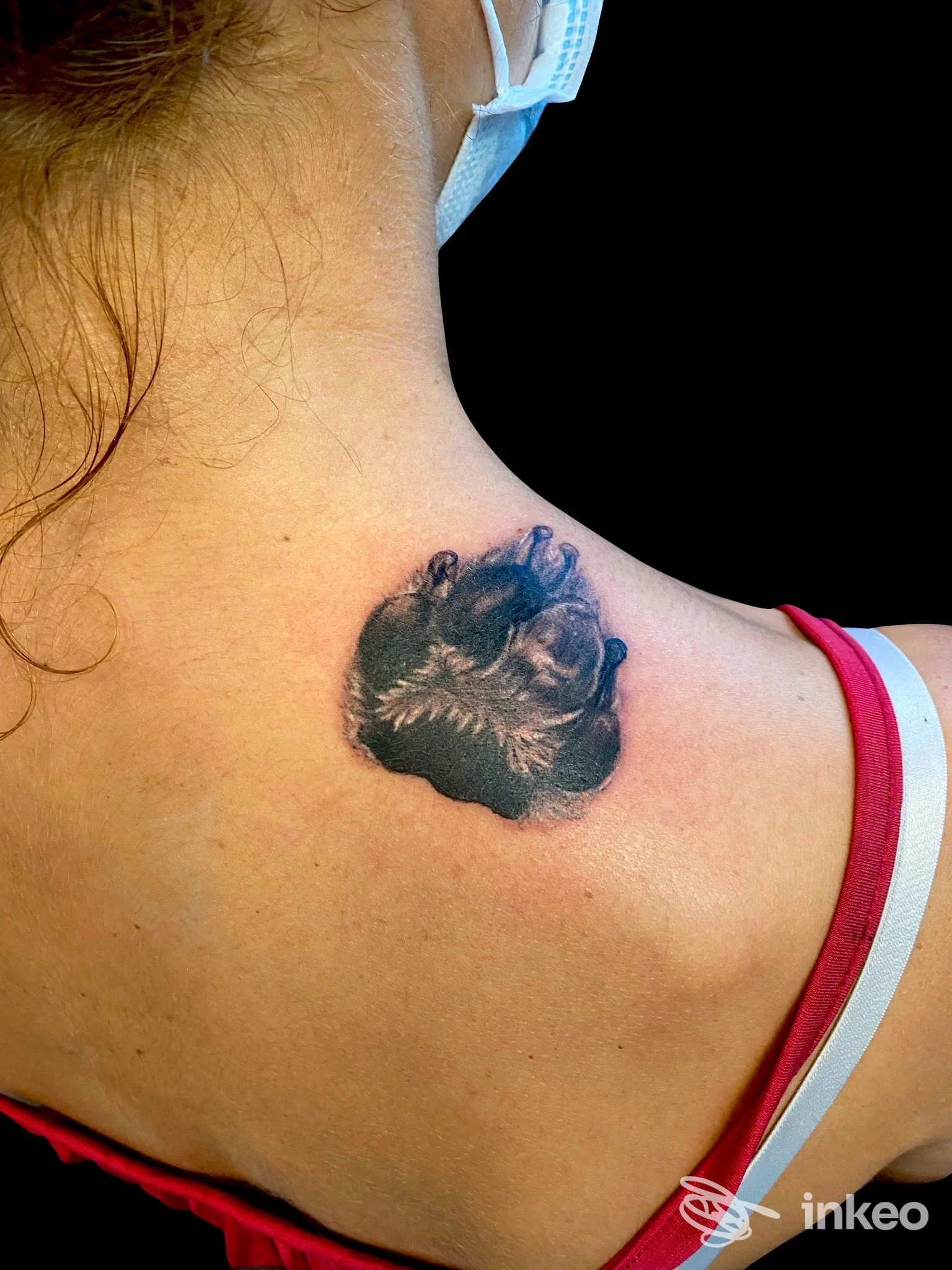 Tattoo nero a forma di fiore stilizzato sulla nuca, visibile sulla pelle, dietro la spalla sinistra della persona.