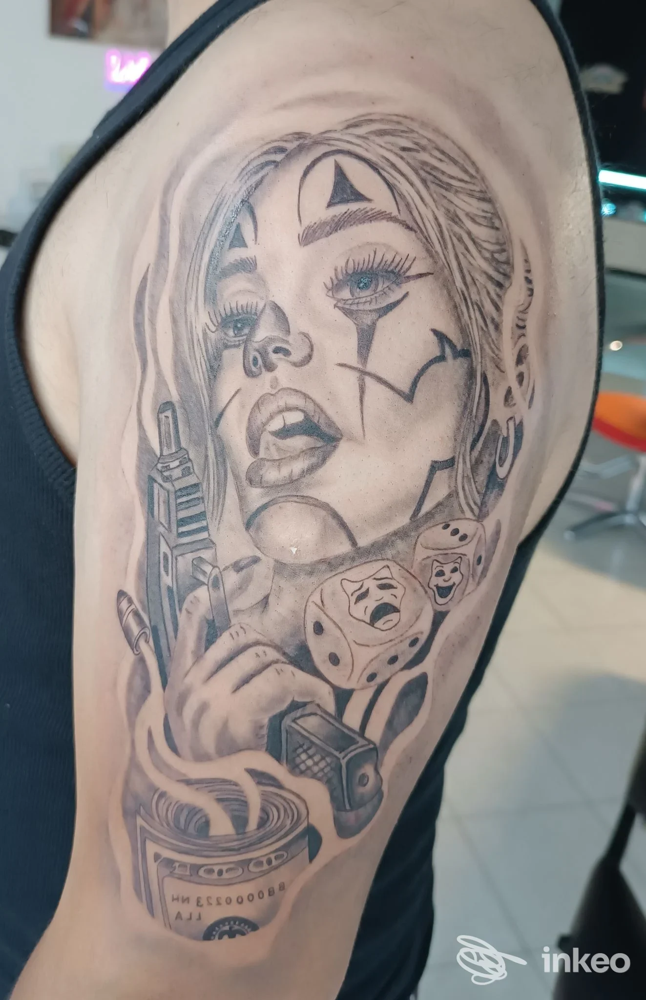 Tattoo visibile sulla spalla: volto femminile stilizzato, pistola, dadi e simboli geometrici, realizzato su pelle umana.