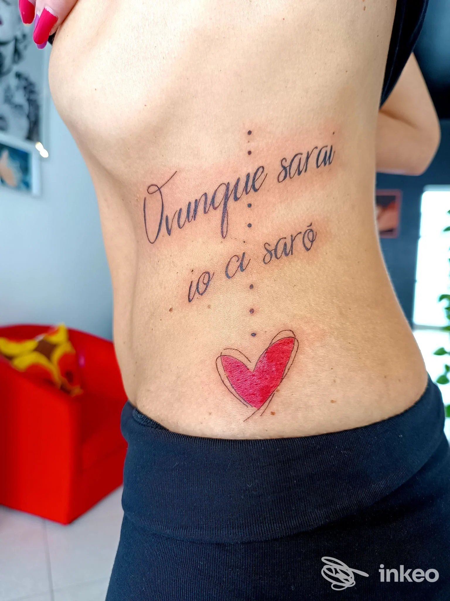 Tattoo sul fianco sinistro con testo corsivo «Ovunque sarò io ci sarò» e cuore rosa, visibile su pelle umana in primo piano, sfondo domestico. Studio Cromatica Tattoo.
