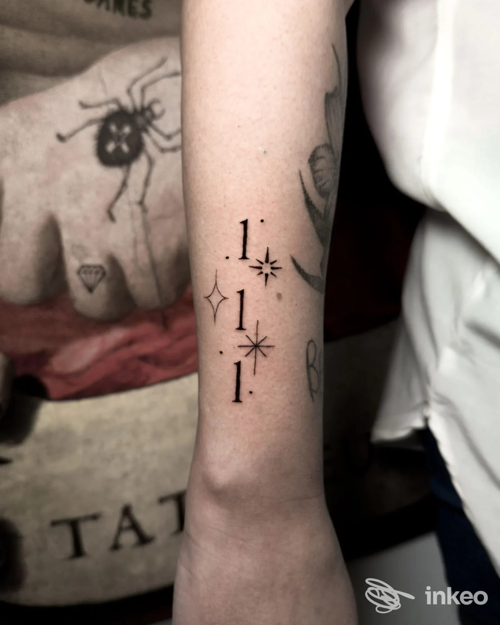 Un tattoo sull’avambraccio: una serie verticale di tre numeri 1 intervallati da piccole stelle, eseguito con linee nette e in nero, senza testo o elementi promozionali visibili.